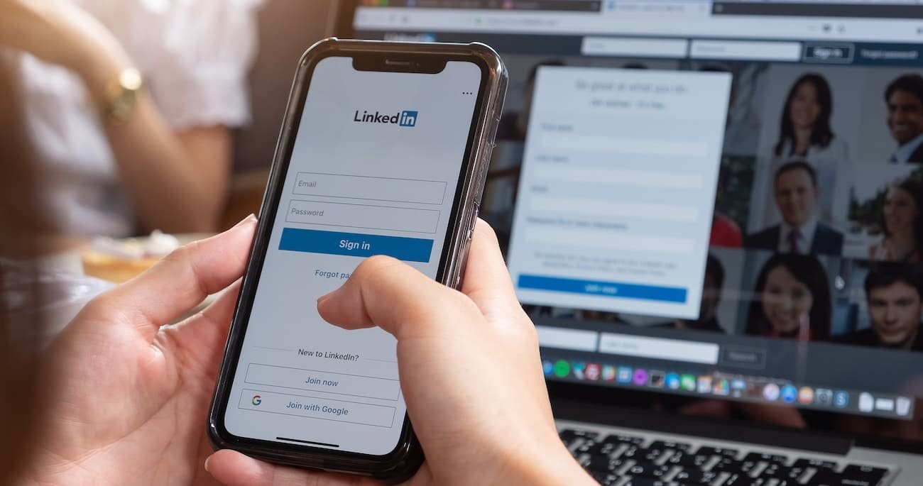 Gestão da rede social LinkedIn: mãos a segurar um telemóvel com o ecrã de entrada do LinkedIn, com um portátil ao fundo